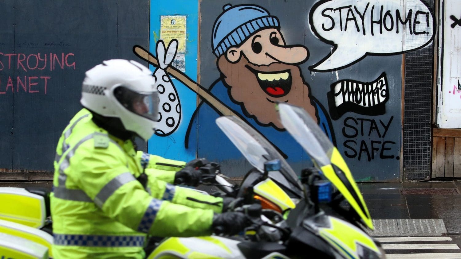 A police officer on a motorbike in front of a mural that advises people 'stay home'