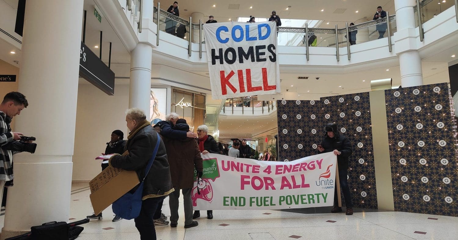 A Fuel Poverty Action Warm Up in a shopping centre. A large banner is hanging from the first floor. It reads "Cold homes kill". A second banner on the ground floor reads "unite 4 energy for all". People are walking around, while a person with a video camera films