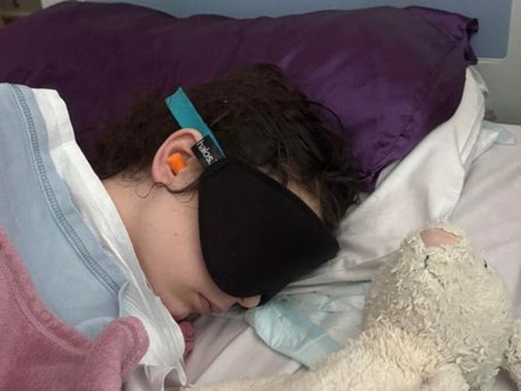 Millie McAinsh lying in bed wearing ear protectors and an eye mask with a teddy lying next to her ME/CFS