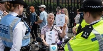 Disabled man Neil Goodwin protesting in his mobility scooter at the entrance to parliament. He is surrounded by police officers and holding signs that read "disabled people against climate change" and ""I cannot run from a climate emergency"