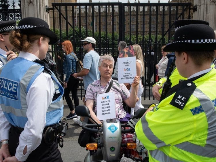Disabled man Neil Goodwin protesting in his mobility scooter at the entrance to parliament. He is surrounded by police officers and holding signs that read "disabled people against climate change" and ""I cannot run from a climate emergency"
