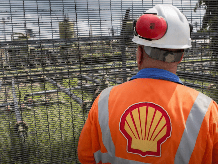 Shell worker with tabard emblazoned with Shell's logo looks out through a fence at oil and gas infrastructure Nigeria