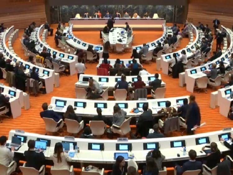 UNCRPD forum room at Geneva there are circles of long tables with people sitting at screens at the top of the room is a long table against a marble stone backdrop