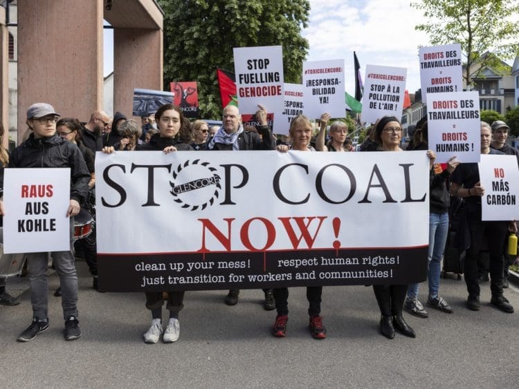 Protesters outside Glencore's AGM gather with a large banner that reads: Stop coal now! Clean up your mess! Respect human rights! Just transition for workers and communities! Others carry placards. One in English reads: Stop fueling genocide Israel