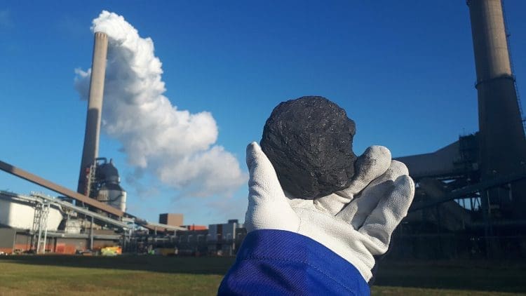 Image shows piece of coal being held in front of coal power plant