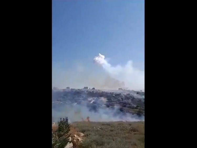 Israel bombing Lebanon with white phosphorus. White smoke and flames rise from an area of grassy hills and trees, with a few houses interspersed between World Environment Day ecocide