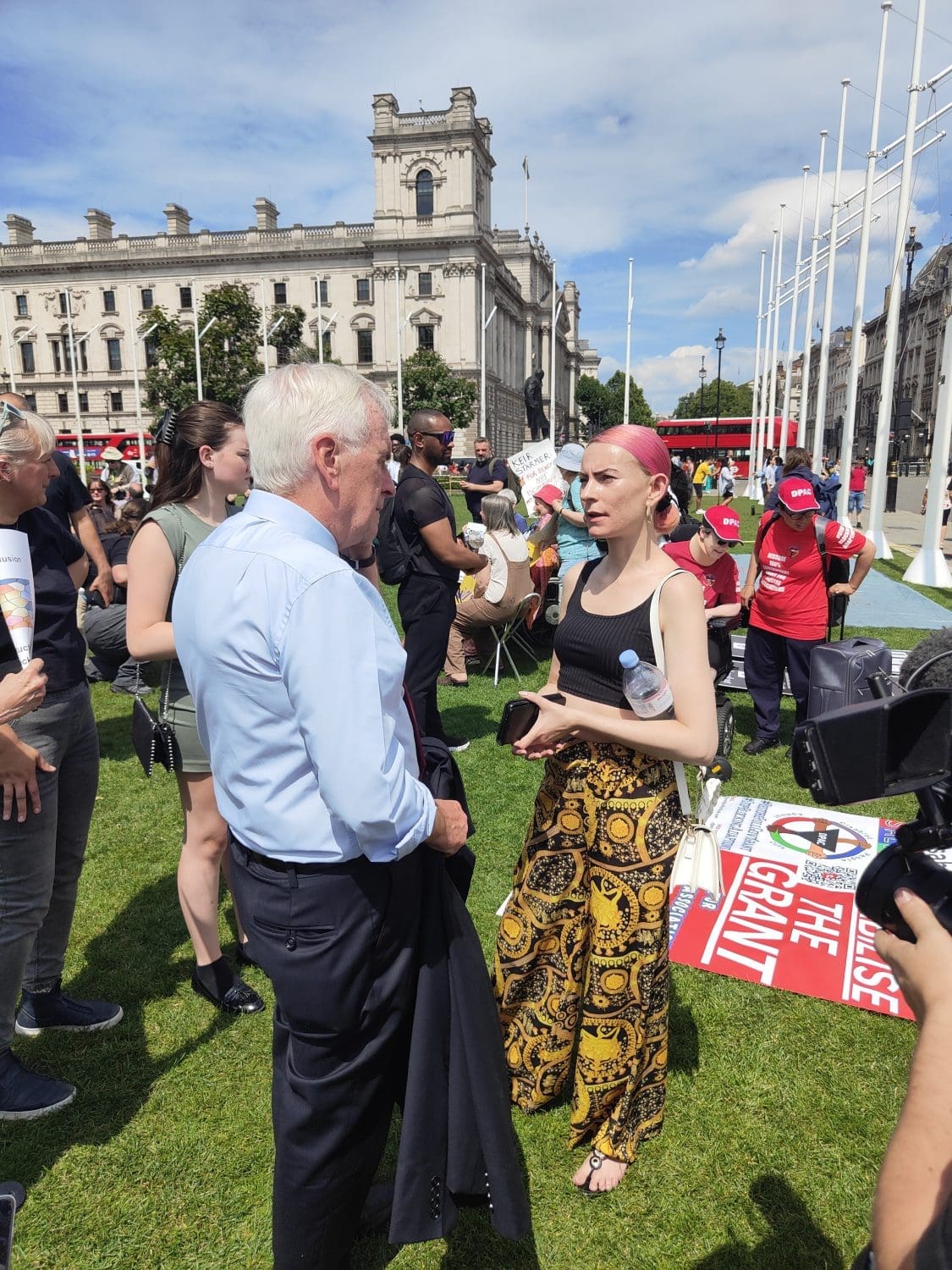 John McDonnell speaking to Nicola Jeffery. He is wearing a shirt and trousers, she has bright pink hair and is wearing a black top and patterned flared trousers disabled people