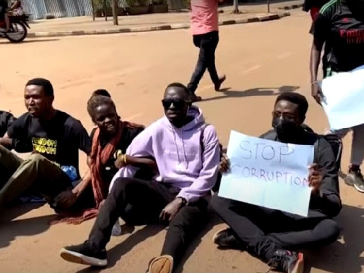 Ugandan anti-corruption protesters sit on the ground with placards.