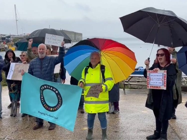 Protesters stand in the rain at Portland port with placards that read: "no floating prison" and "asylum process - EU: 6-9 months - UK: 1-3 years - stuck in limbo". bibby Stockholm Labour