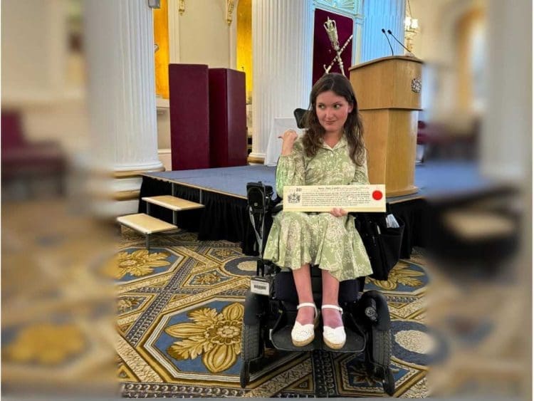 Anna Landre in her wheelchair pointing at a stage which only has steps leading up to it. She is holding her Freedom of the City of London award, in an old fashioned room with large white pillars and patterned carpet. Anna is wearing a green patterned dress and white shoes and she looks sad